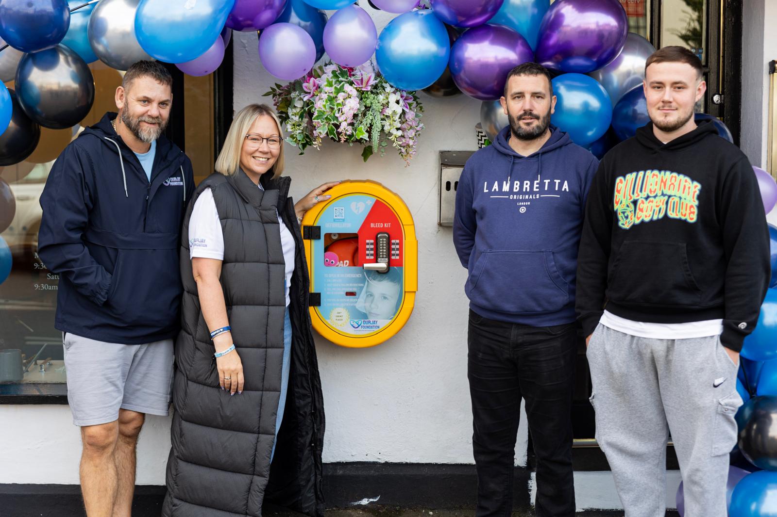 People gathered beneath a balloon arch at the unveiling of a community defibrillator cabinet.