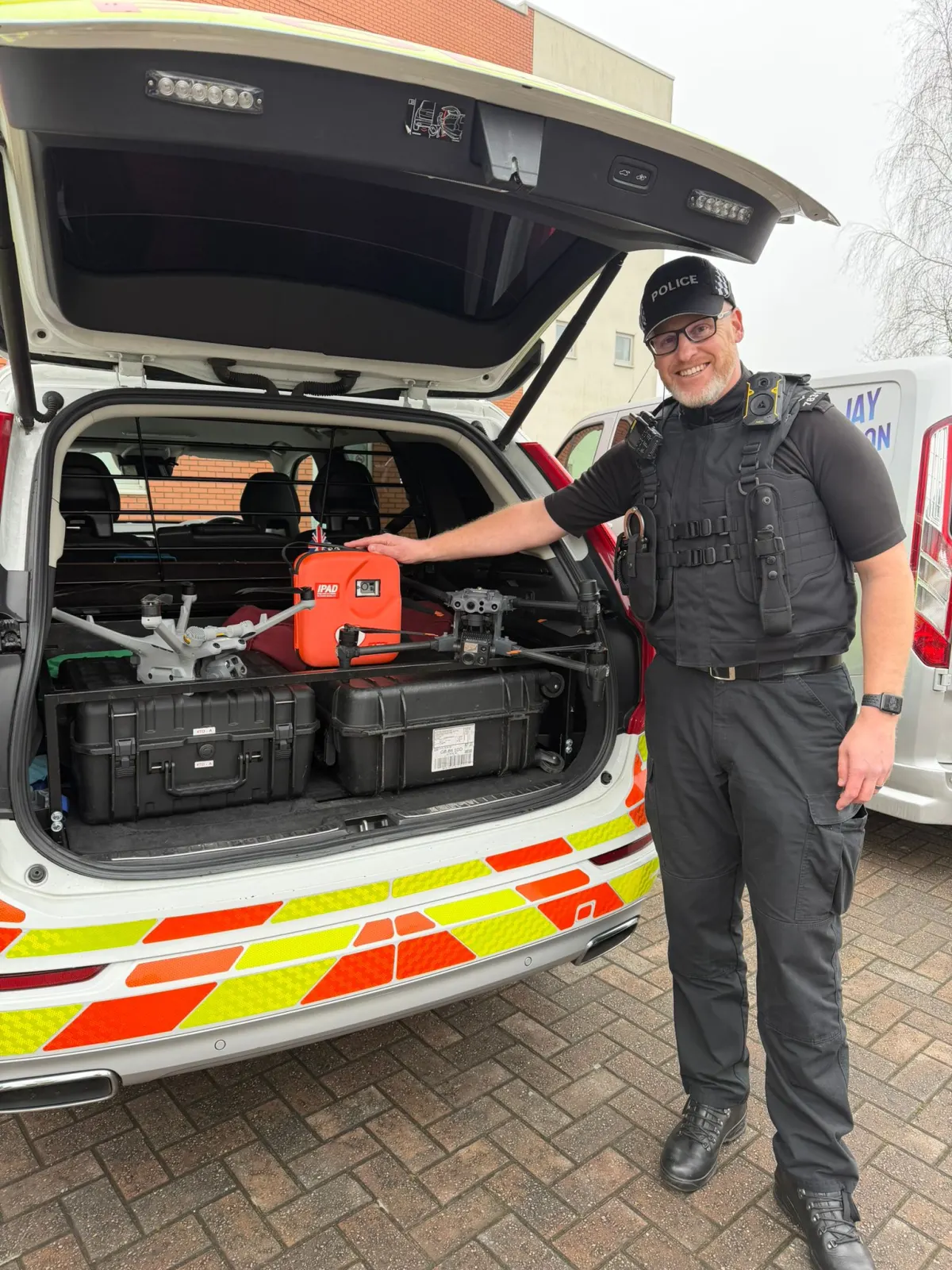 A specialist response officer in body armour stands at the open boot of an unmarked car containing equipment cases and an orange AED.