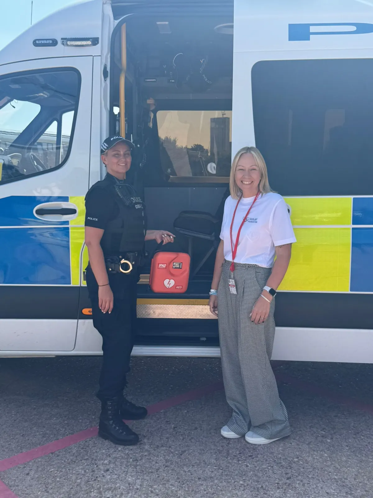 A police officer and a supporter wearing an OurJay Foundation t-shirt stand together in front of an open police van, holding an iPAD AED unit between them.