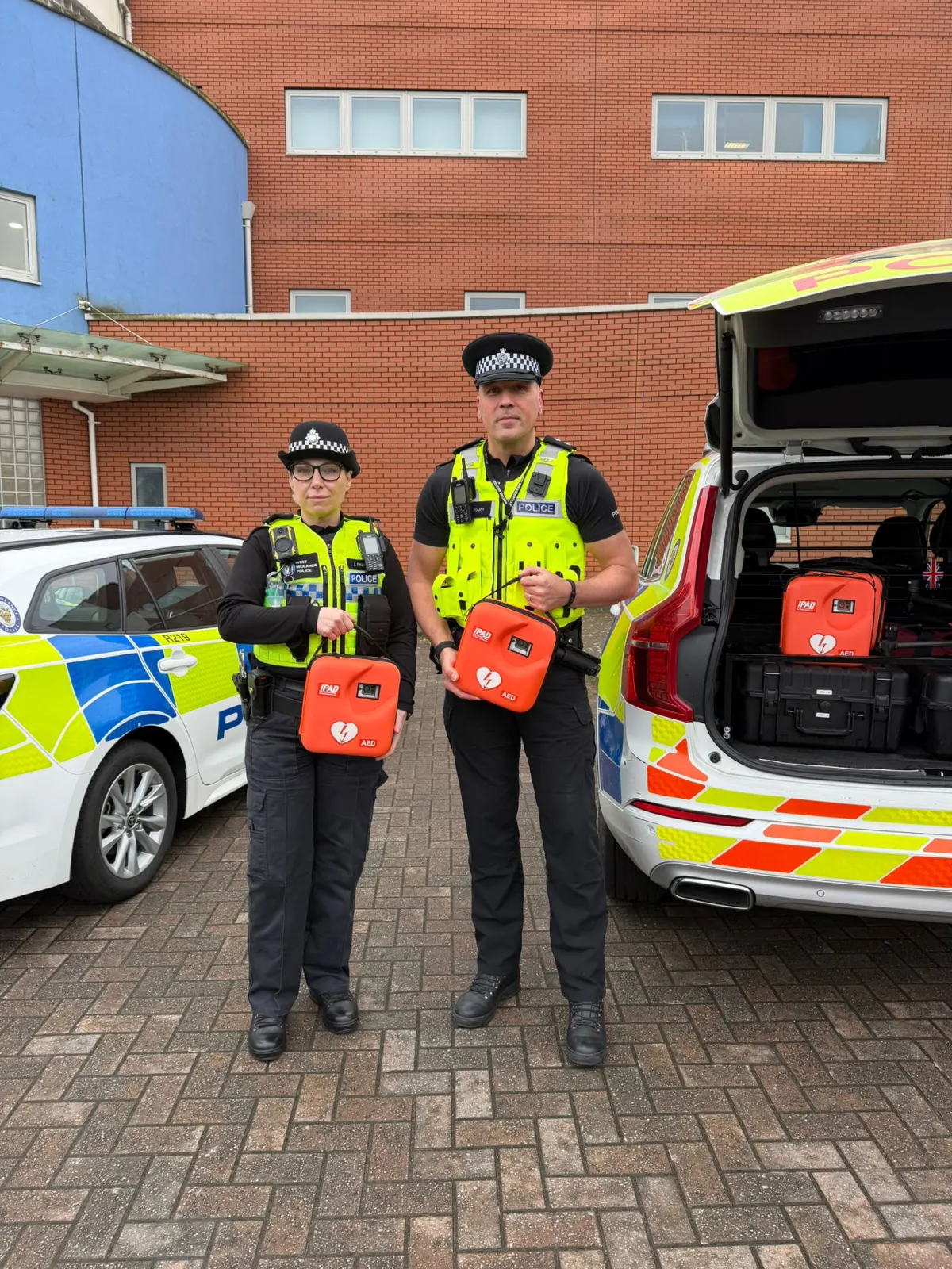 Two uniformed police officers standing in front of marked patrol vehicles, each holding an orange iPAD AED unit.
