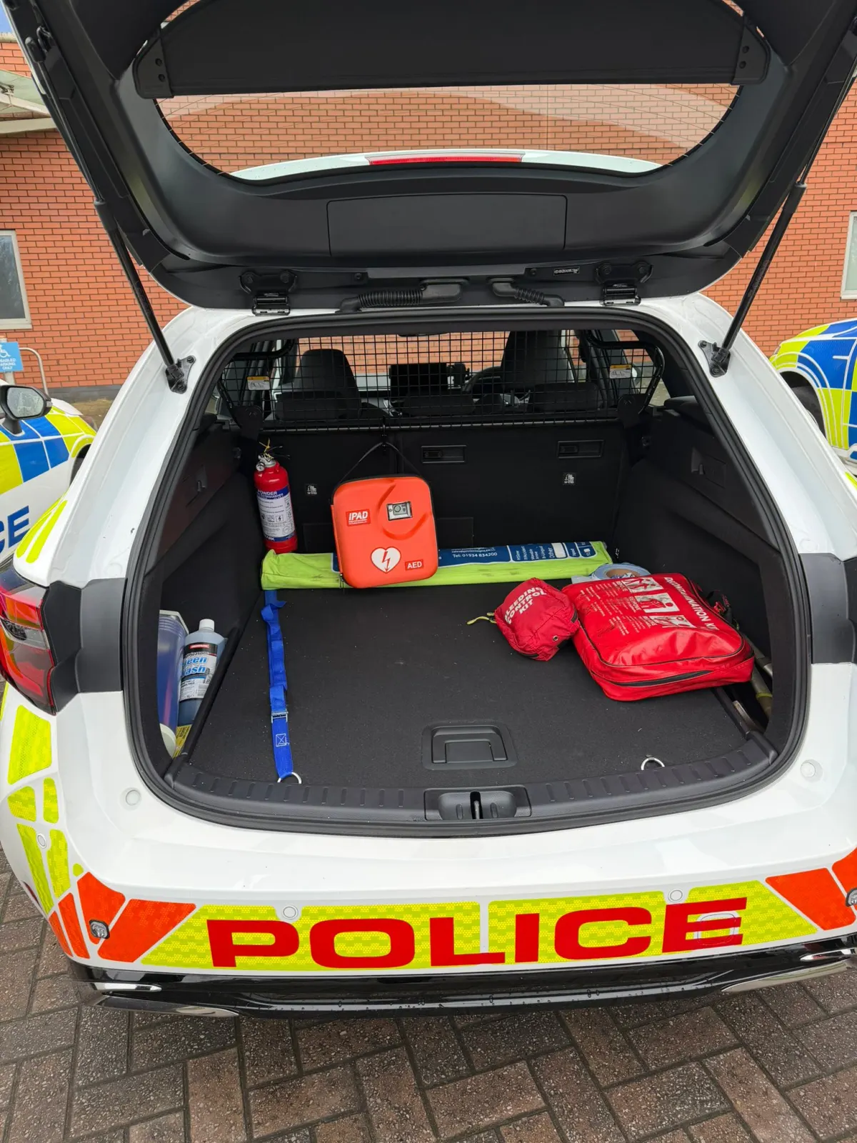 Open boot of a marked police patrol car showing an iPAD AED, a red trauma kit, and a fire extinguisher loaded for response.