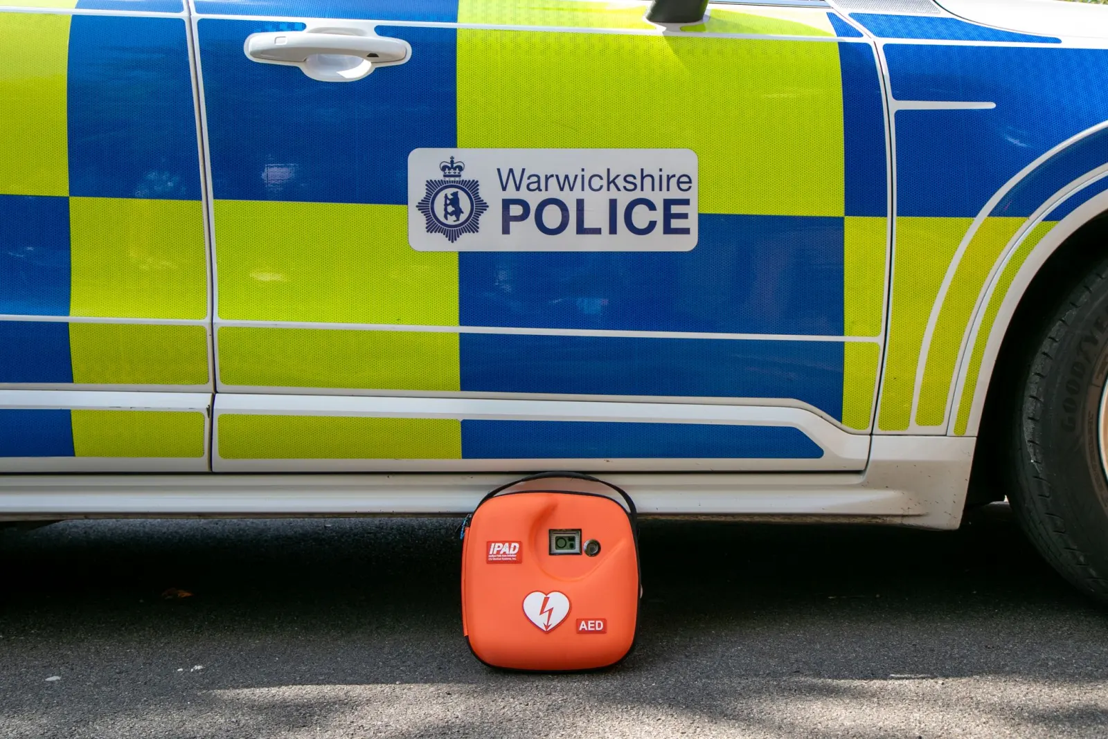 A Warwickshire Police marked patrol vehicle with an orange iPAD AED unit on the ground beside it.