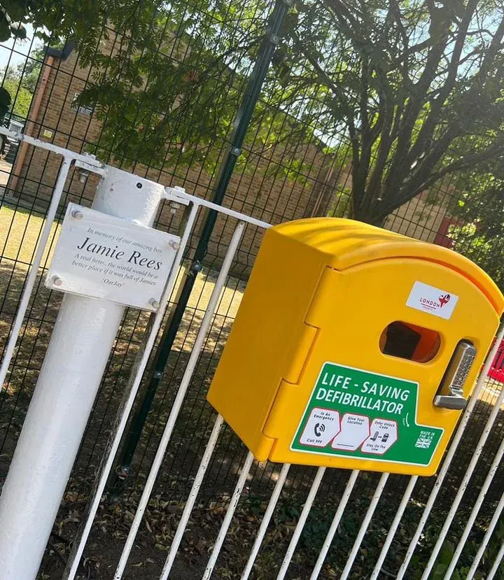 Yellow public-access defibrillator cabinet mounted on a green school railing with a 'Jamie Rees' name plaque.