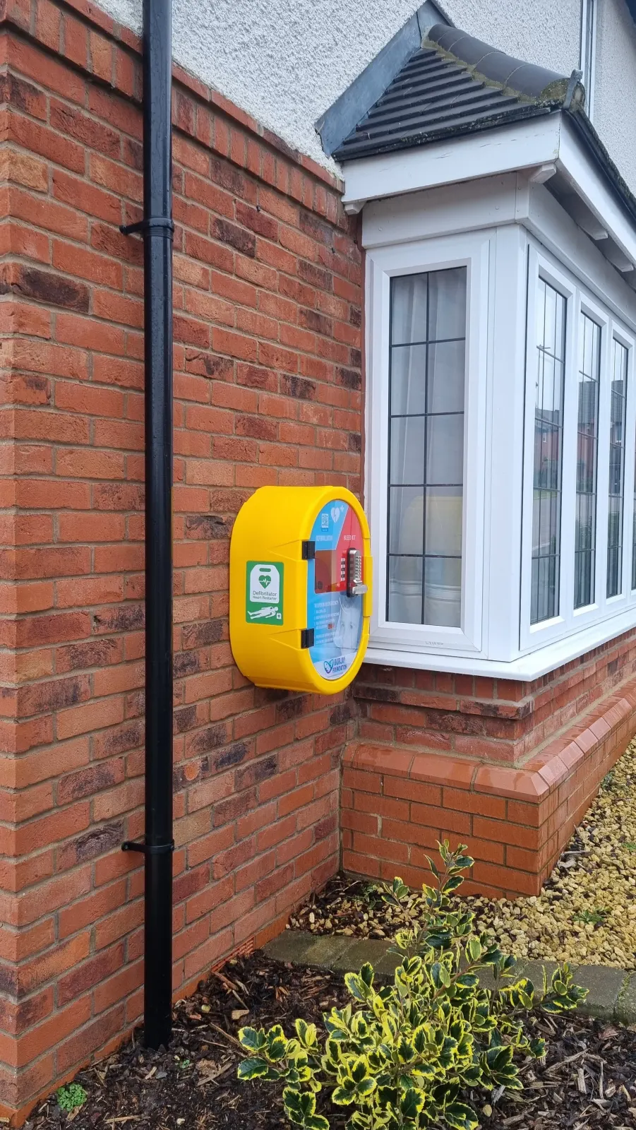 A yellow public-access defibrillator cabinet mounted on the brick wall of a residential house.